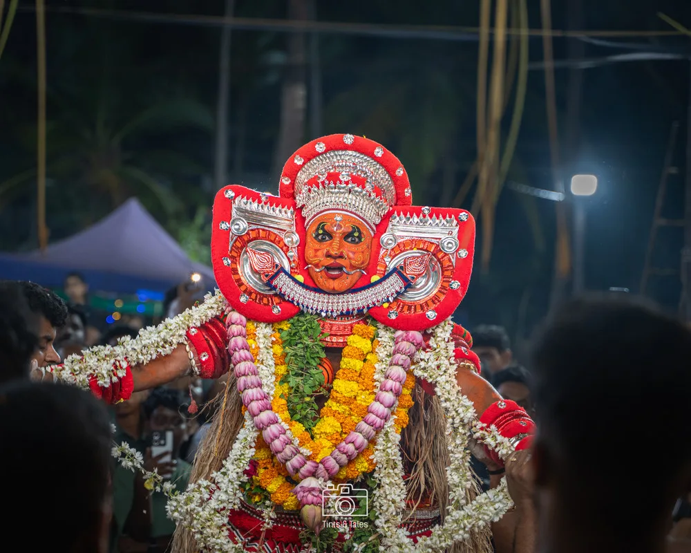 A striking photograph of a Theyyam ritual performer in Kerala, wearing elaborate traditional costume and heavy face paint.